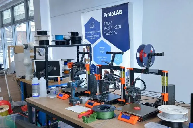 Trzy drukarki 3D i szpule filamentu na stole warsztatowym, w tle plansza ProtoLAB